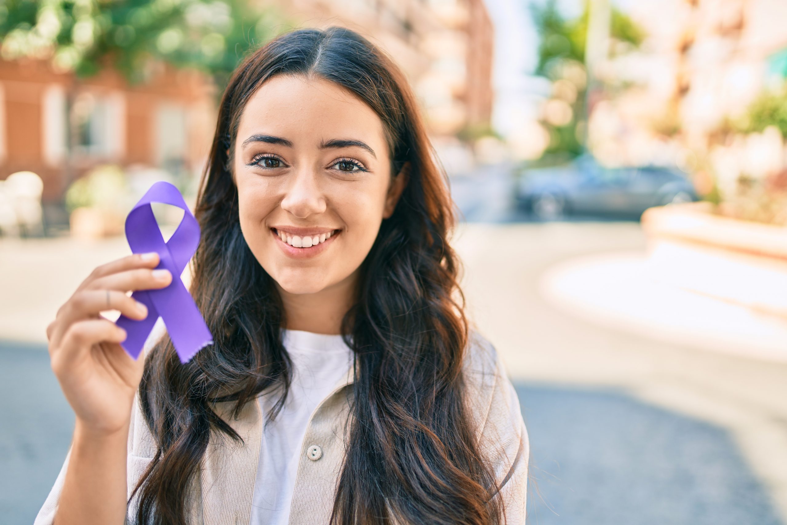 Young hispanic woman smiling happy holding purple ribbon walking at the city.